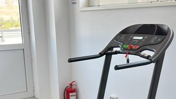 Una cinta de correr en una habitación con un tapete de ejercicios azul, un extintor y una ventana.
