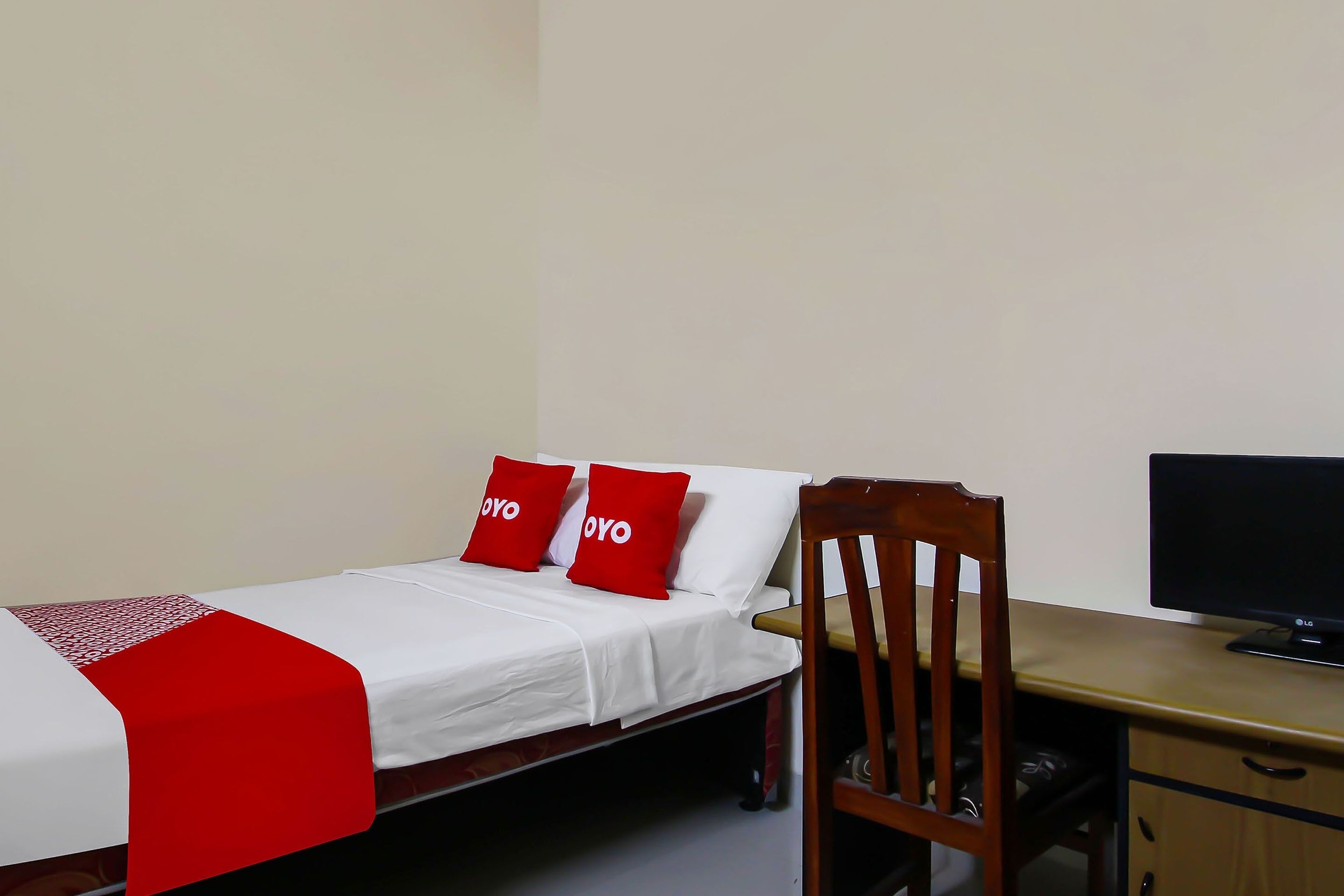 A single bed with white bedding and red pillows, a wooden chair, a desk with a computer, and a wardrobe.