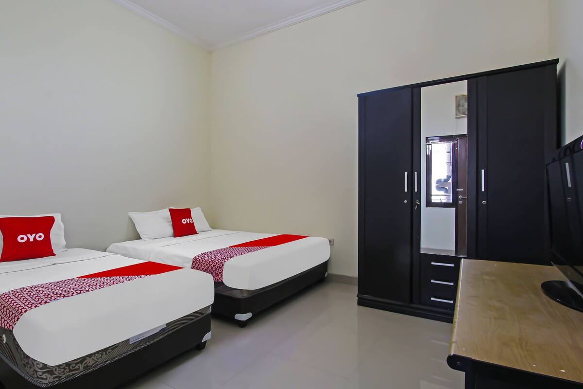 Habitación de hotel con dos camas, cada una con una colcha y almohadas rojas y blancas, un escritorio de madera y un armario negro.