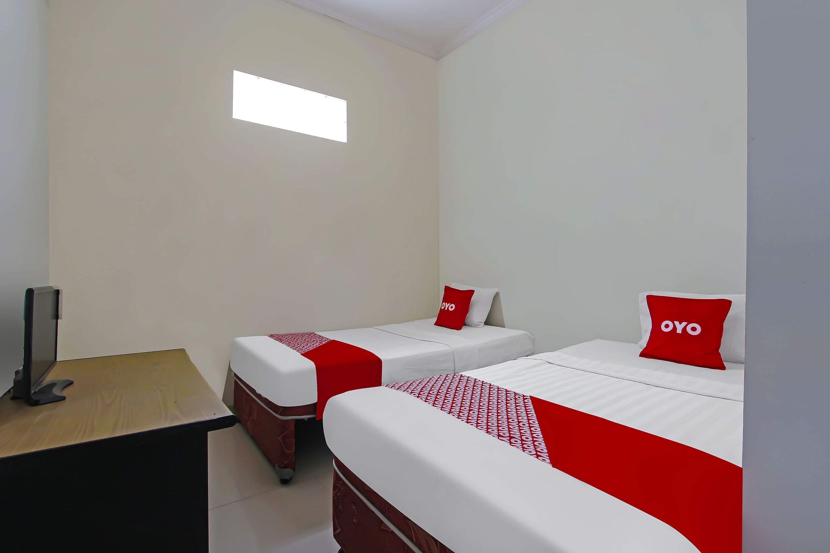Habitación de hotel con dos camas, cada una con colcha y almohadas rojas y blancas, un escritorio con computadora y un ventanal que proporciona luz natural.