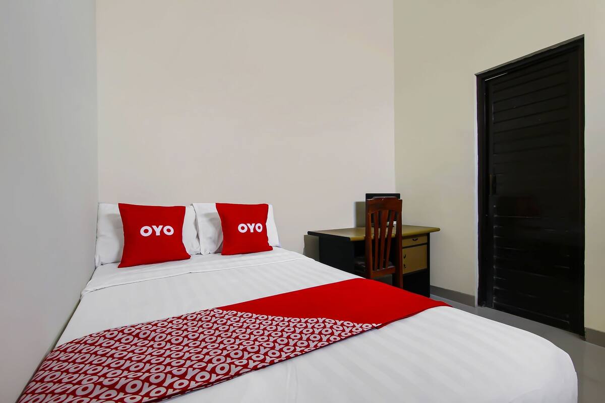 Una cama bien hecha con ropa de cama blanca y detalles rojos, un pequeño escritorio de madera y una puerta corrediza negra.