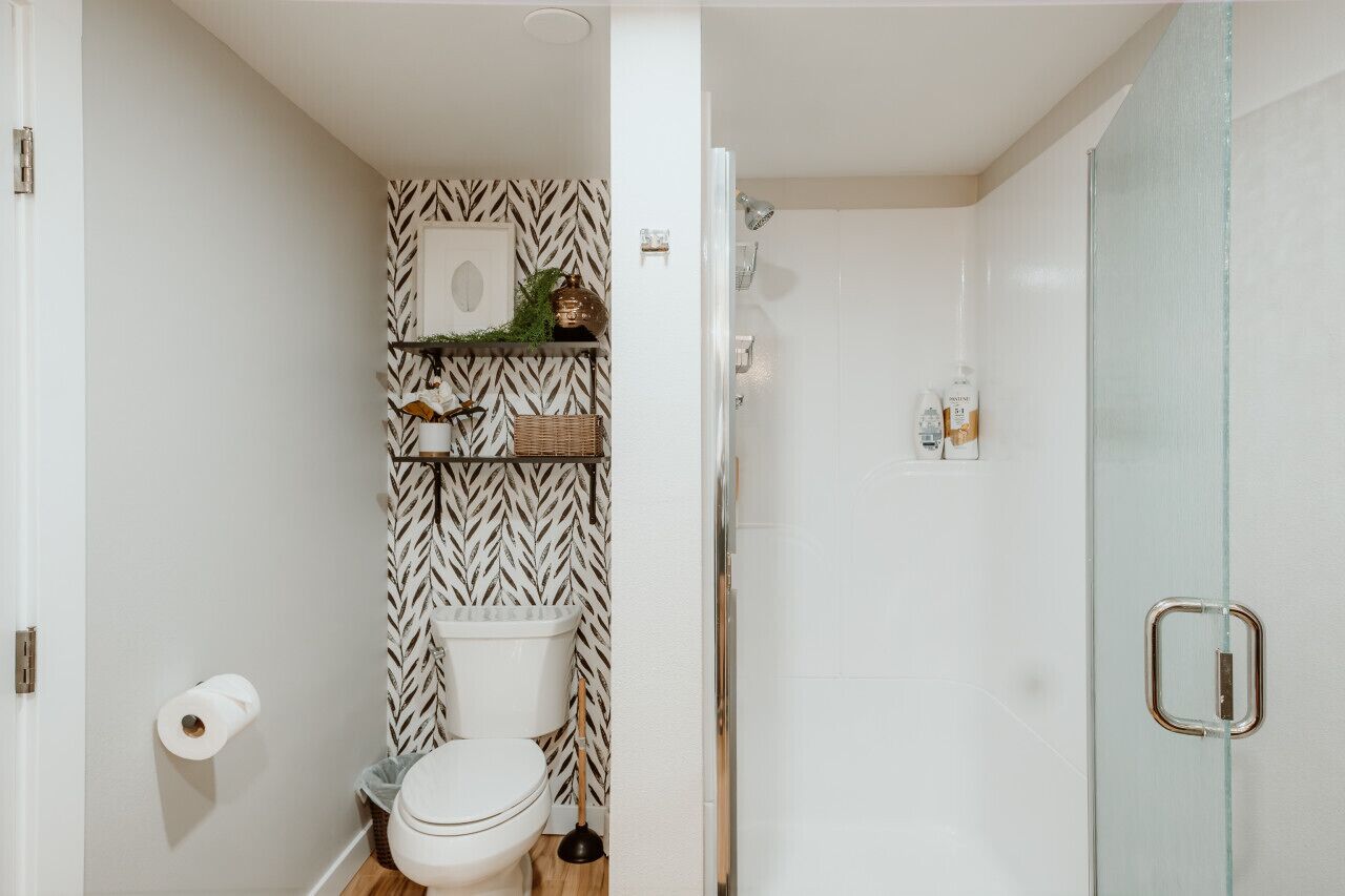 A compact bathroom with a white and black patterned wall, a toilet, a shelf with toiletries, and a glass shower door.