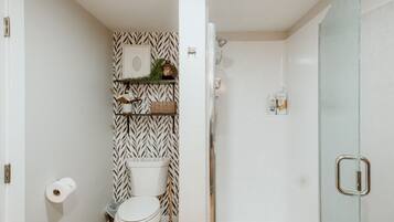 A compact bathroom with a white and black patterned wall, a toilet, a shelf with toiletries, and a glass shower door.