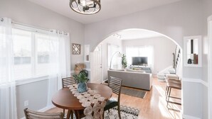 Un comedor con una mesa redonda, sillas y una araña. Se aprecia una sala de estar al fondo con un sofá y un televisor.