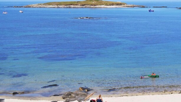 Una playa con agua azul cristalina, una orilla arenosa y una pequeña isla a lo lejos.