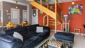 Una sala de estar moderna con un sofá negro, una mesa de centro de vidrio y un juego de ajedrez. Una escalera de madera conduce a un nivel superior.