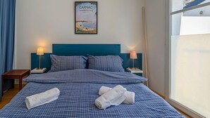 Ein Schlafzimmer mit einem Bett, zwei Nachttischen, einem Beistelltisch, einem gerahmten Bild an der Wand und einem Fenster mit Blick auf Gebäude.