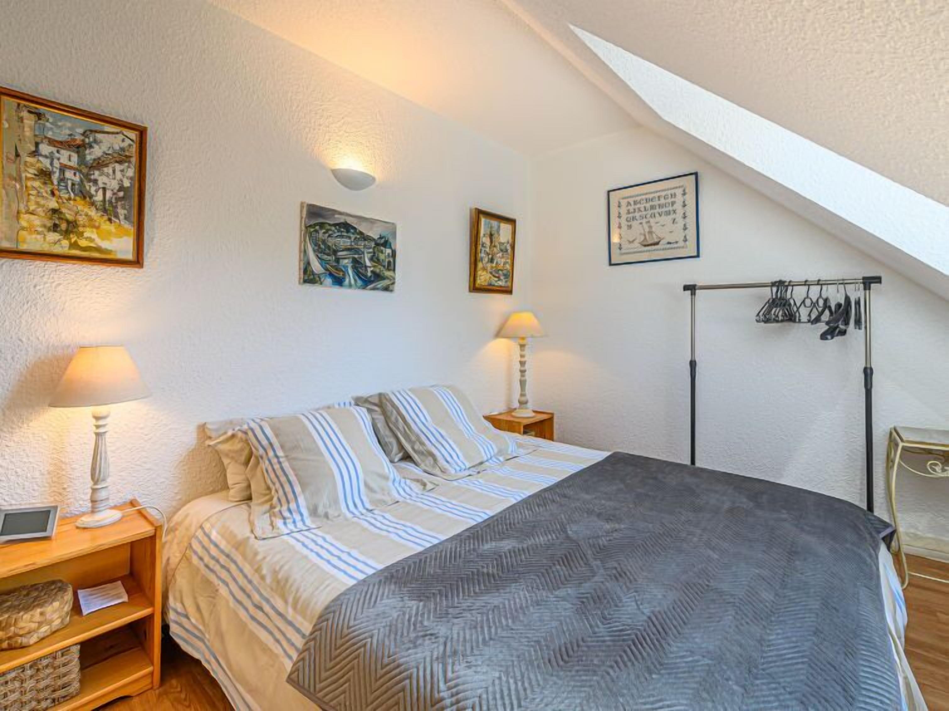 A bedroom with a bed, two bedside lamps, a small bedside table, a coat rack with clothes, and a framed picture on the wall.