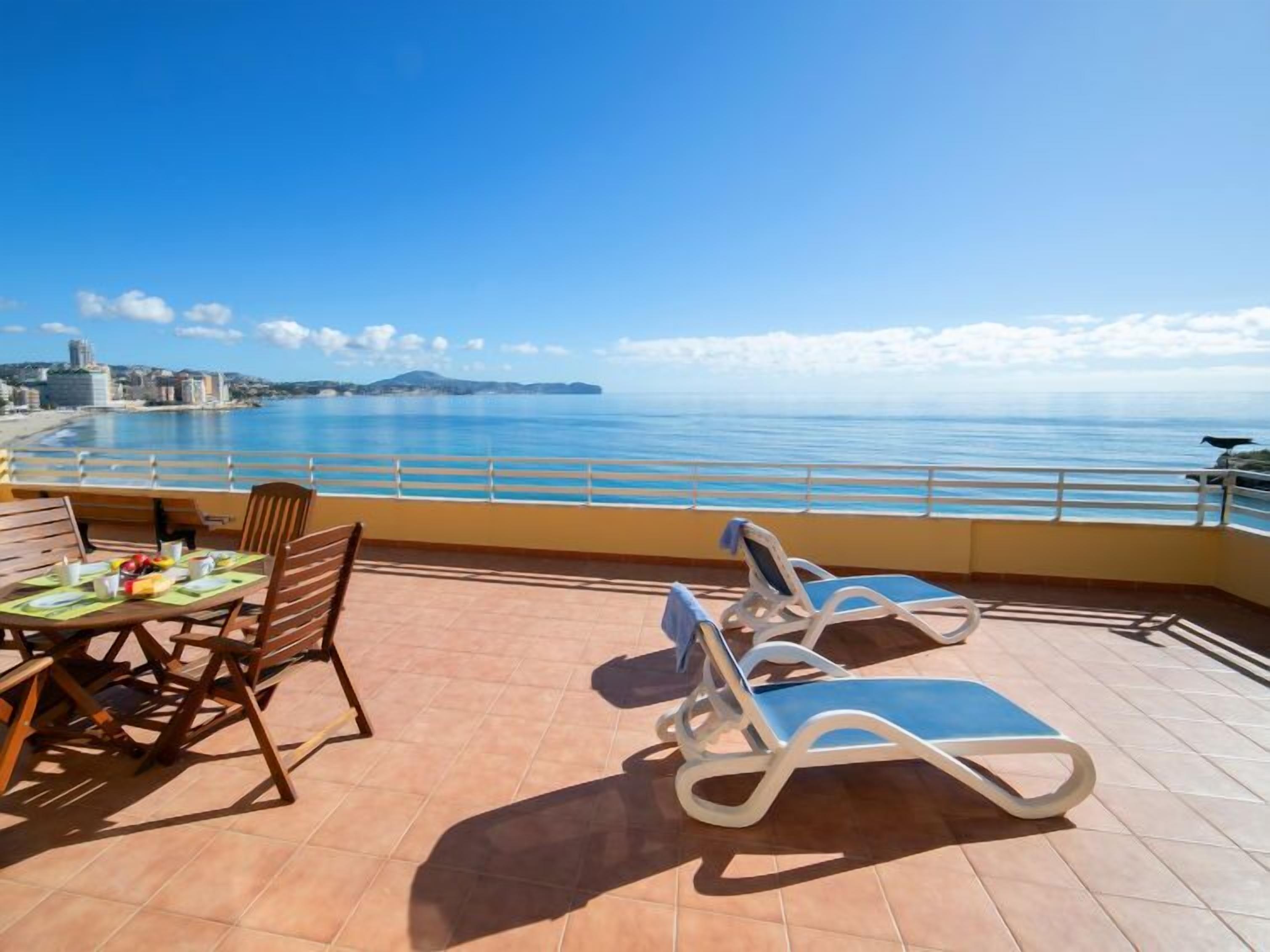 Eine Terrasse mit Esstisch, zwei Liegestühlen und Blick auf das Meer und die Stadt.