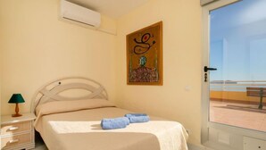 Quarto com cama, mesa de cabeceira, luminária, pintura e vista para a praia através de uma porta de vidro.