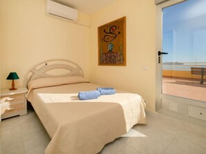 Une chambre à coucher avec un lit, une table de chevet, une lampe, un tableau et une vue sur la plage, visible à travers une porte vitrée.