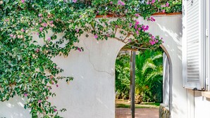 Un bâtiment blanc, doté d’un socle en pierre et d’une arche recouverte de feuillage vert et de fleurs roses.