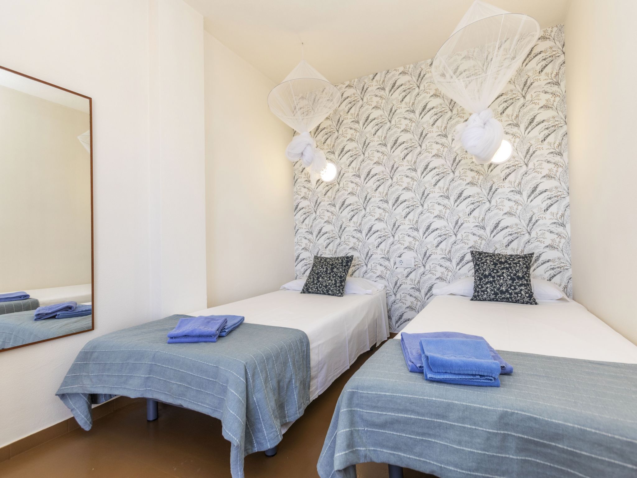 A hotel room with two single beds, each with a blue-striped bedspread and a white pillow. The room features a large mirror on one wall and a patterned wallpaper with a leaf design on the opposite wall.