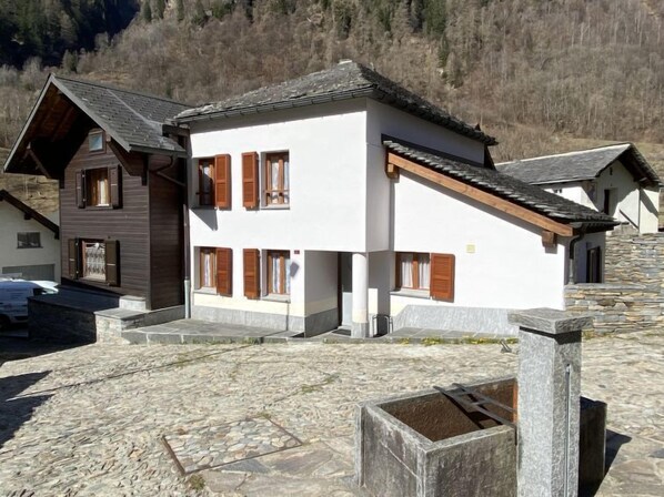 Un bâtiment à deux étages, doté d’une façade blanche et de volets en bois, situé dans une région montagneuse où les rues sont pavées de pierres.