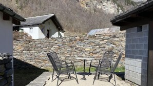 A stone wall with a rustic building in the background, a stone table with metal chairs, and a clear view of the surrounding mountains.