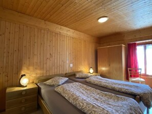 Ein Holzhütten-Zimmer mit zwei Betten, einem Nachttisch, einem Kleiderschrank und einem Fenster mit roten Vorhängen.