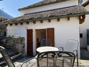 Ein kleines Gebäude mit einer Steinmauer, einem Ziegeldach und einer Tür mit Holzklappläden. Draußen stehen Metallstühle und ein Tisch.
