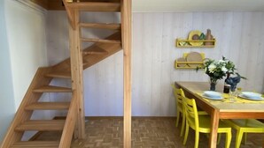 A dining area with a wooden staircase, a wooden table, yellow chairs, and a white wall with shelves. - Della Posta Stambecco by Interhome (Rossa)