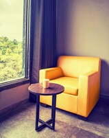 Una habitación con un sillón amarillo, una mesita redonda con una taza blanca y una ventana con cortinas.