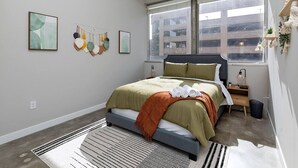Un dormitorio moderno con una cama grande, una ventana con vistas a edificios, una mesita de noche pequeña y una estantería con plantas.