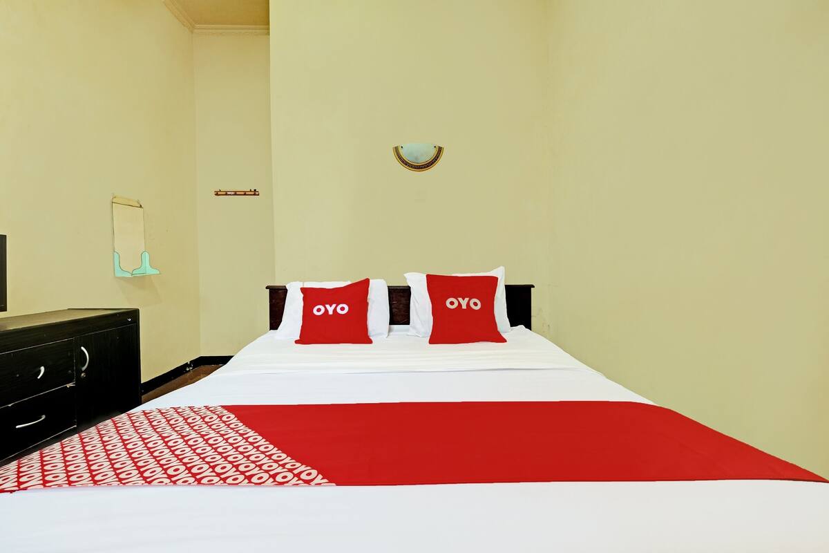 A hotel room with a bed, two red pillows with "OYO" text, a red and white patterned throw, a wooden dresser, and a wall-mounted light fixture.