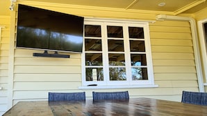 A covered patio with a wooden table and chairs, a large flat-screen TV mounted above a window, and a view of trees through the window.