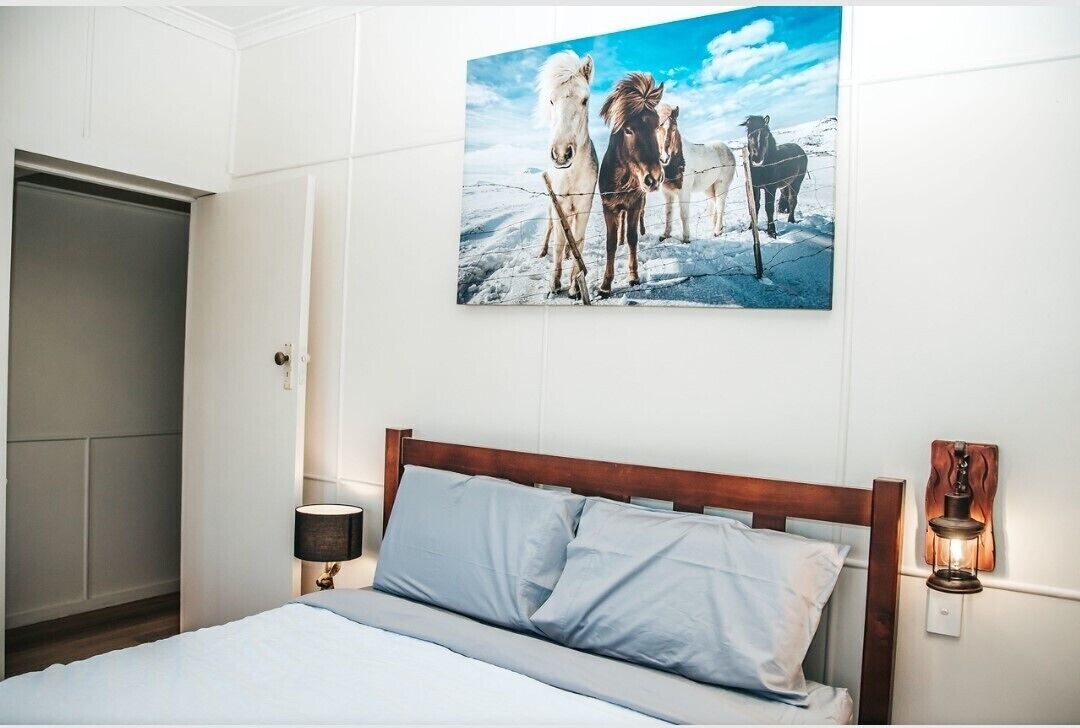 A bedroom with a bed, a nightstand, a wall-mounted lamp, and a painting of horses.
