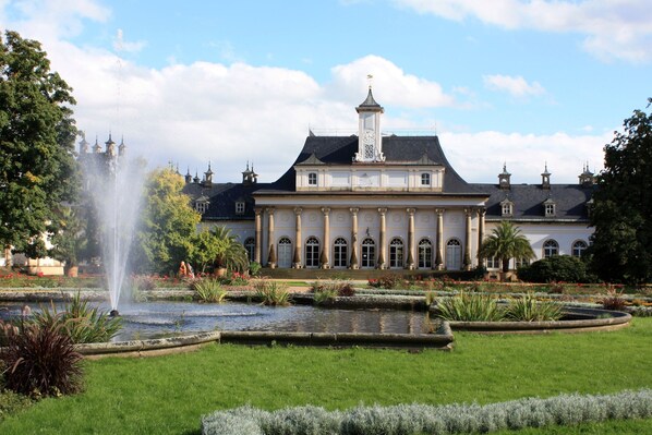 A grand building with a fountain in the foreground, surrounded by well-maintained gardens and a clear sky.