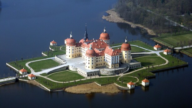 Ein großes, historisches Gebäude mit mehreren Kuppelbauten, umgeben von Wasser und Grünpflanzen.