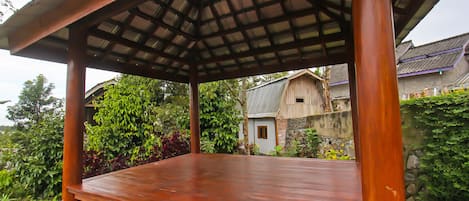 Un pabellón de madera con techo inclinado, rodeado de abundante vegetación y un muro de piedra.