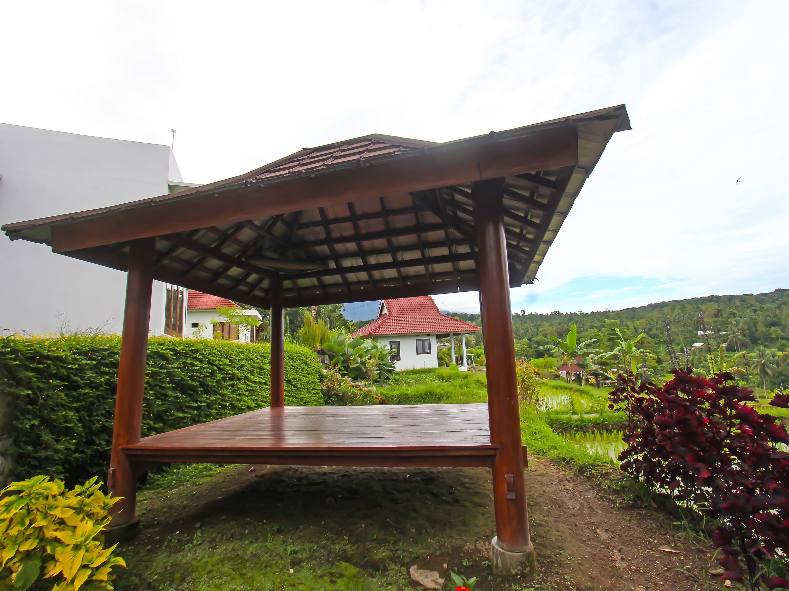 A wooden gazebo with a sloped roof, surrounded by lush greenery and a view of houses in the background.