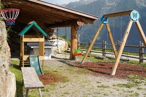 A wooden play structure with a swing set and a slide, a picnic table, and a building with a sloped roof. - Chalet Ulten (Santa Valburga)