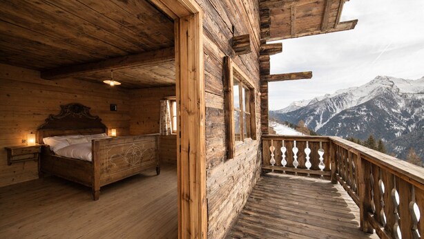 Eine Holzhütte mit einem Schlafzimmer, das ein Bett, einen Nachttisch und ein Fenster beinhaltet. Der Raum verfügt über einen Holzbalkon mit Geländer und Blick auf schneebedeckte Berge.