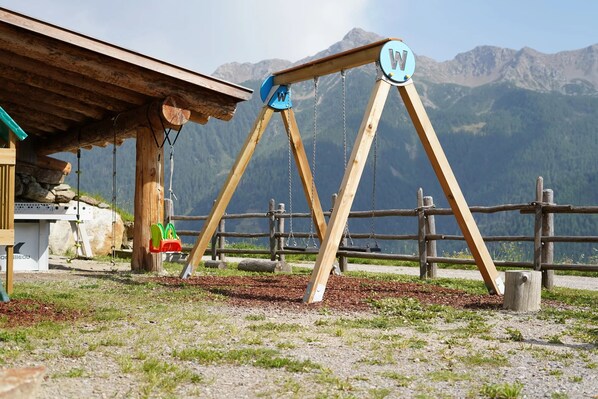 Un ensemble de jeux d’extérieur en bois, comprenant une balançoire en forme de « W » et de « V », une structure en bois et un paysage de montagne en arrière-plan.