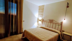 A bedroom with a bed, curtains, a stone wall, and a nightstand with a lamp. - MASSERIA TENUTA SOFIA (Otranto)