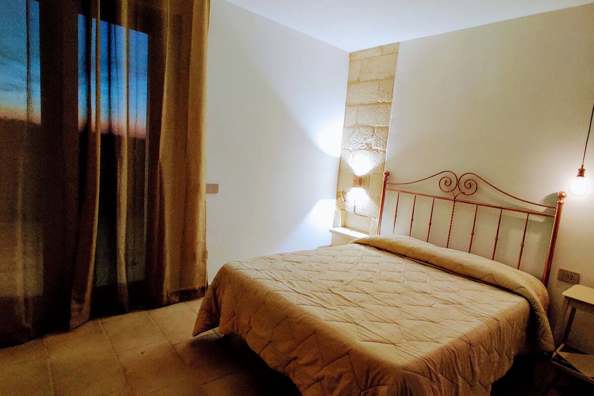 A bedroom with a bed, curtains, a stone wall, and a nightstand with a lamp.
