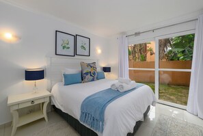 A bedroom with a bed, two bedside lamps, a nightstand, a window with curtains, and two framed botanical prints on the wall.