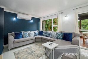 A modern living room with a grey sectional sofa, patterned cushions, a small white side table, and a view of greenery through the window.
