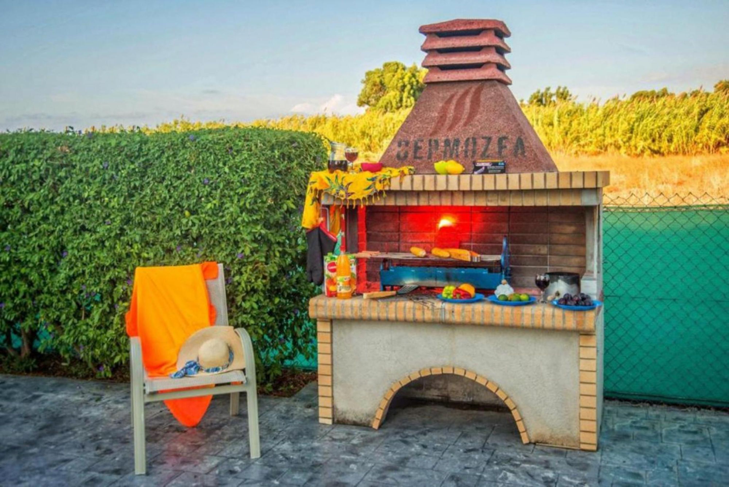 Ein Backofen aus Ziegelsteinen mit der Aufschrift 'PIZZA', umgeben von einem Zaun und einem Stuhl mit einem orangefarbenen Stoff.