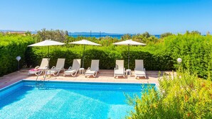 A poolside area with lounge chairs and umbrellas, surrounded by greenery and a clear view of the sea.