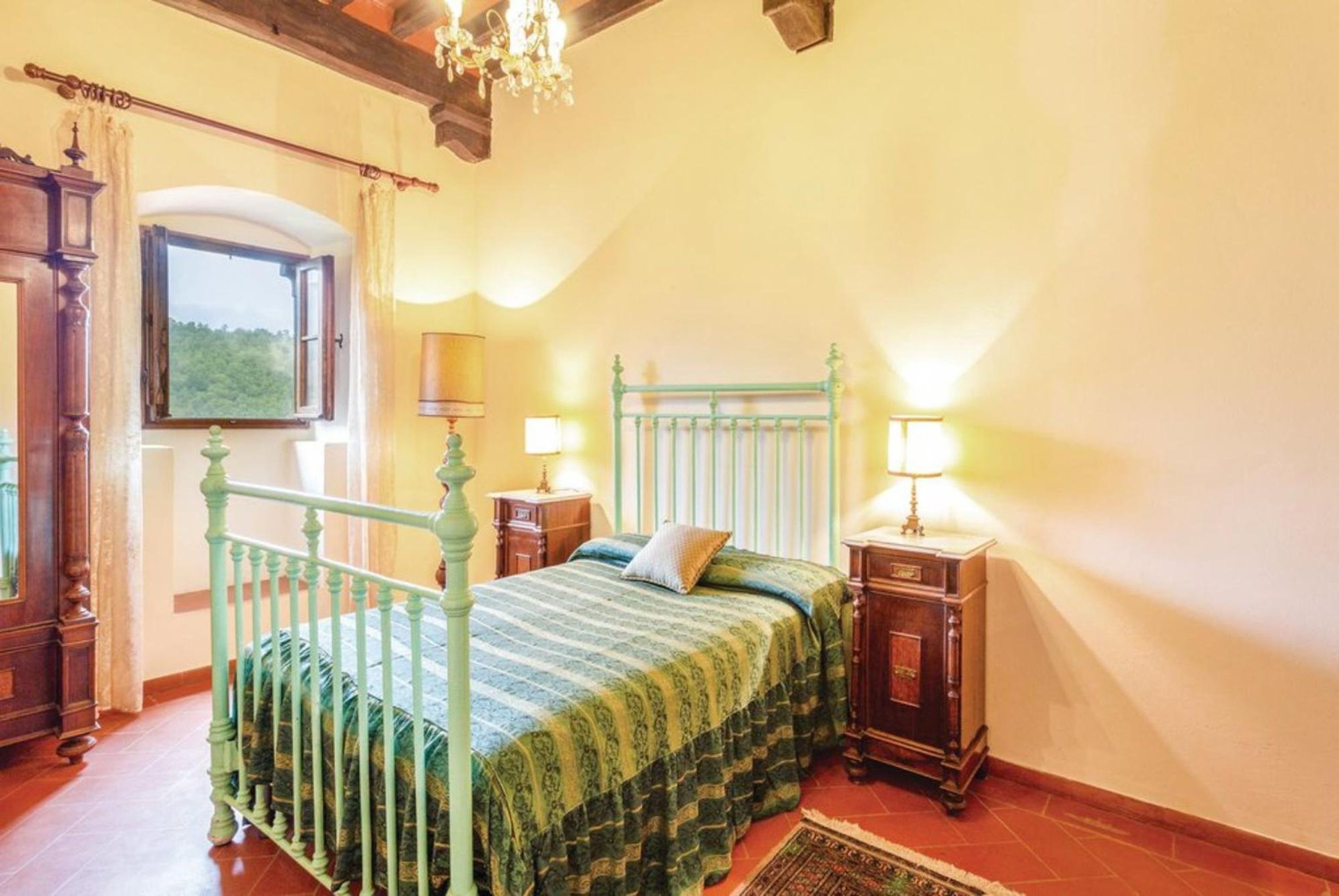 A bedroom with a bed, a nightstand, a lamp, a window, and a chandelier.