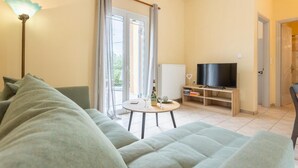 Ein Wohnzimmer mit Sofa, Fernseher, einem kleinen Tisch mit Flasche und Glas sowie einem Fenster mit Vorhängen.