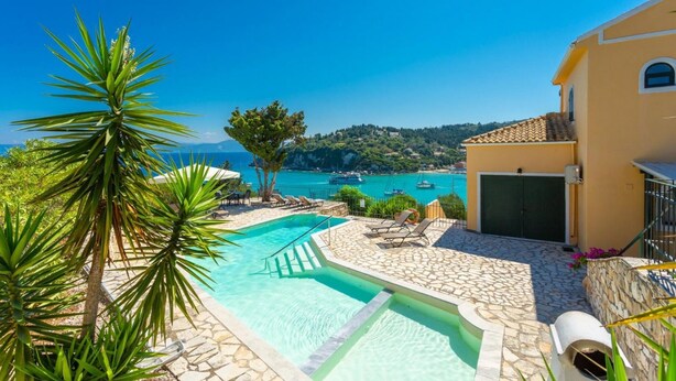 A pool area with clear blue water, a stone deck, and a view of boats on the sea. - Lakka Bay View Collection (Paxos)