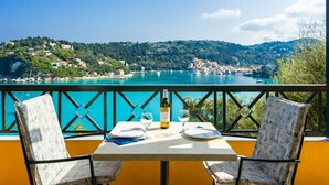 A table with two chairs on a balcony overlooking a coastal town with a clear view of the sea and hills. - Lakka Bay View Collection (Paxos)