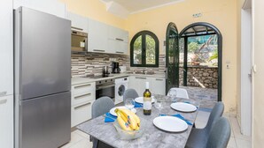 A compact kitchen with modern appliances, a dining area with a table set for two, and a view of a garden through an arched doorway. - Lakka Bay View Collection (Paxos)