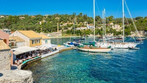 A coastal marina with boats docked, a stone building with outdoor seating, and a hillside with houses in the background. - Lakka Bay View Collection (Paxos)