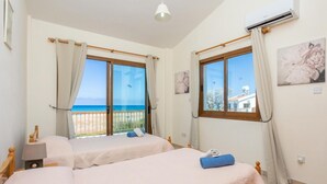 A room with two beds, a window with a view of the sea, and an air conditioning unit. - Blue Bay Villas Collection (Polis)
