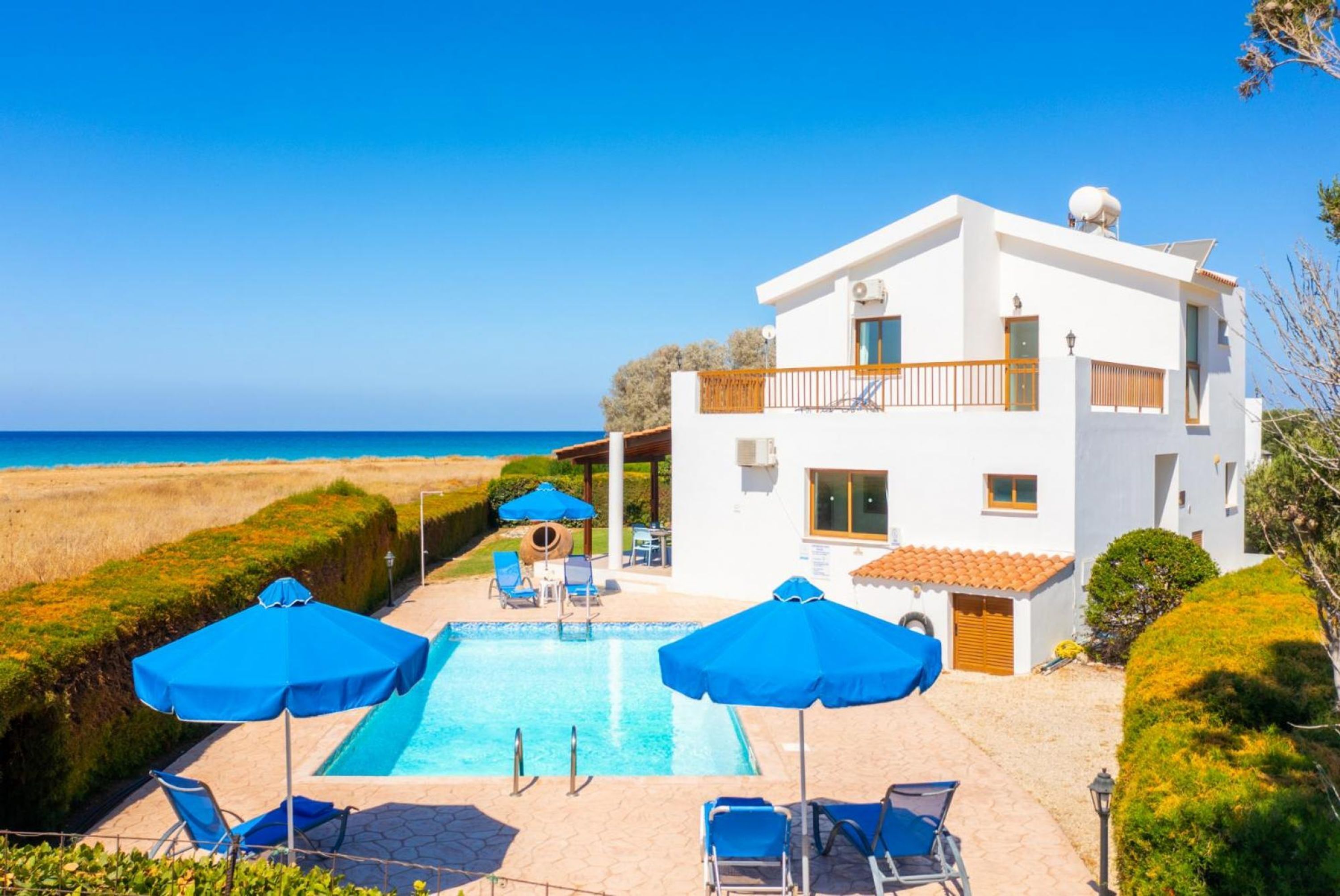 A two-story white building with a pool, blue umbrellas, and a view of the sea.