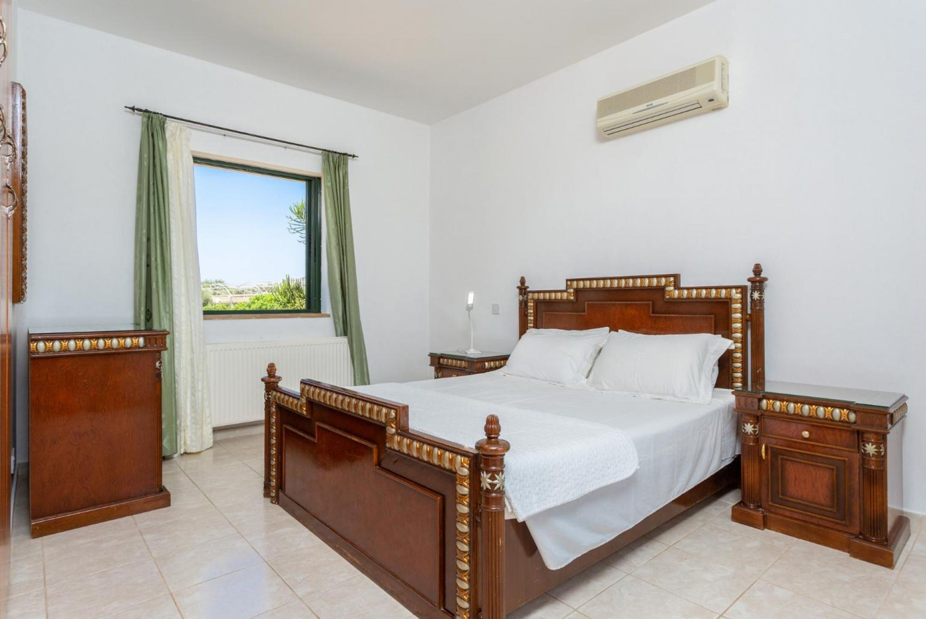 A bedroom with a wooden bed, a nightstand, a dresser, a window with curtains, and an air conditioning unit on the wall.