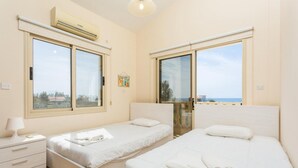 A hotel room with two single beds, a bedside table with a lamp, and a large window offering a view of the sea. - Rose Villas Collection (Ayia Marina Chrysochous)